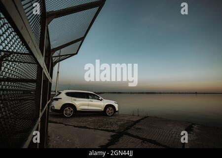 White compact SUV car with sport and modern design parked on concrete road by the sea near the beach at sunset Stock Photo