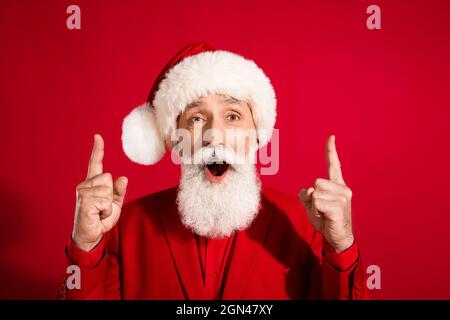 Photo of shocked funky santa claus indicate forefingers up empty space open mouth wear x-mas hat suit on red color background Stock Photo