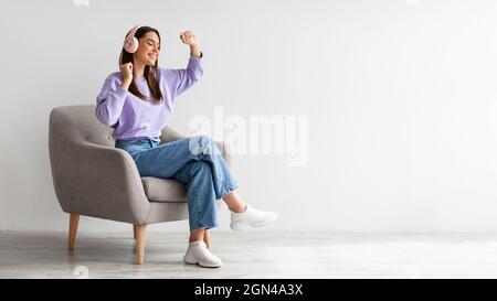 Cheery young lady in headphones listeing to music and dancing while sitting in armchair against white studio wall Stock Photo