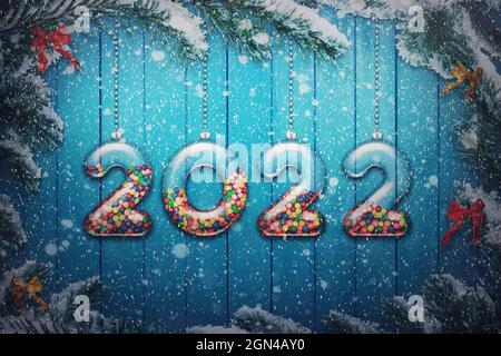 Happy New Year 2022 holiday background. Set of transparent numbers made of glass filled with multi colored candy and sweets hang on snowy Christmas tr Stock Photo