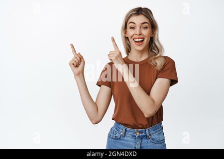 Happy smiling blond girl pointing fingers at upper left corner, showing advertisement, making announcement, standing against white background Stock Photo