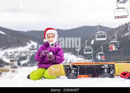 Cute adorable preschooler caucasian kid girl portrait little girl in santa hat and snowboard enjoy winter sport activities. Stock Photo