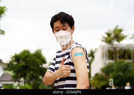 Young Asian man wearing covid-19 face protection mask showing thumbs up gesture at while looking at bandage after vaccine for protection against corona virus during day