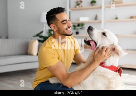 Happy young Arab man stroking his adorable dog in living room. Human animal friendship concept Stock Photo