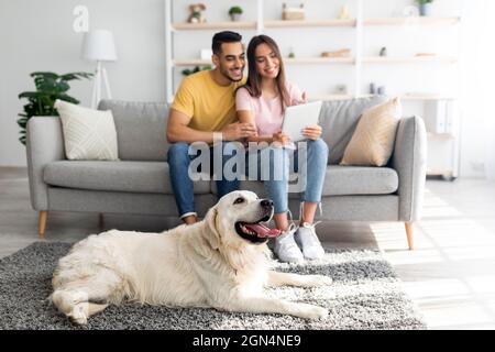 Affectionate diverse couple using tablet on couch, cute pet dog lying on the floor at home, selective focus Stock Photo