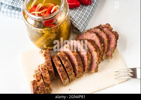 Sliced meatloaf on kitchen board, glass jar with pickled cucumber and vegetable, towel on white background. Stock Photo