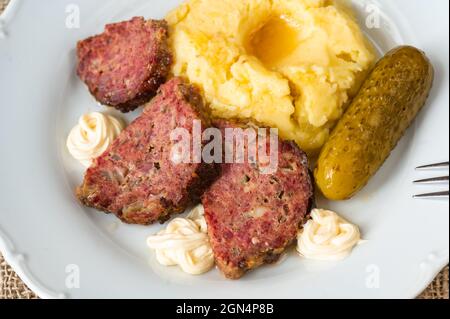 Pieces of baked meatloaf, mashed potato, mayonnaise and pickled cucumber, portion of main meal on white plate, fork, closeup. Stock Photo