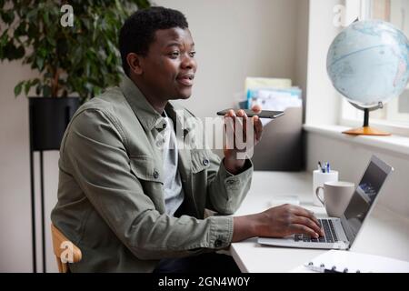 Enthusiastic Young Man Working From Home On Laptop Speaking Into Mic On Mobile Phone Stock Photo