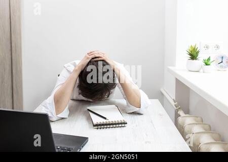 Middle-aged woman with short-haired brunette in glasses works at a laptop near window. The office worker put her head in her hands, bent over the tabl Stock Photo
