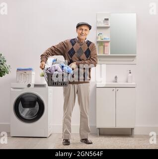 Smiling elderly man holding a laundry basket full of clothes inside a bathroom Stock Photo