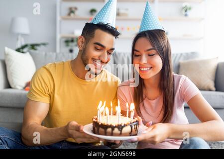 Portrait of happy young multiracial couple in festive hats holding birthday cake with lit candles at home Stock Photo