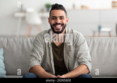 Portrait of handsome young Arab man smiling and looking at camera, sitting on sofa at home Stock Photo