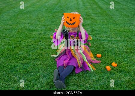 Funny happy little girl in halloween costume is sitting on grass. rick or trunk. Happy Halloween. Stock Photo
