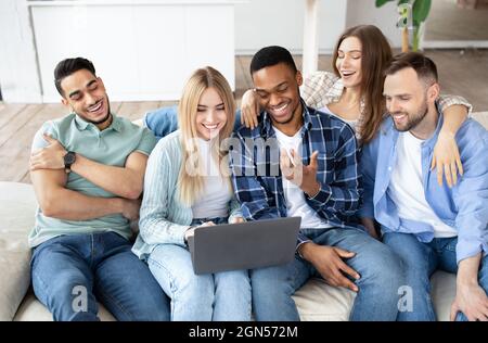 Cheerful diverse friends using laptop pc, sitting on sofa in living room, having video call, chatting online at home Stock Photo