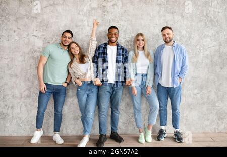 Full length portrait of multiracial young friends posing and smiling at camera, standing against grey studio wall Stock Photo
