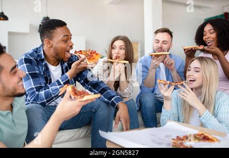 Positive interracial friends having home party, sitting on couch, eating pizza, having fun together indoors Stock Photo