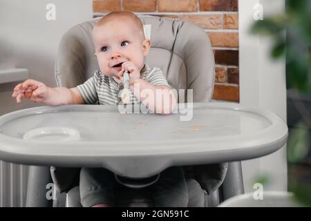 First baby food pure. Mother giving spoon of vegetables or fruits in feeding chair at home. Cute child trying to eat by himself. healthy support Stock Photo