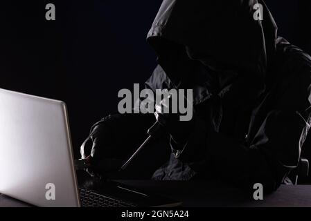 Criminal hacker in a hood in black clothes and balaclava destroys a laptop with tools, a screwdriver and pliers on a black background Stock Photo