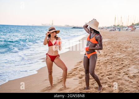 Full body of positive young multiracial girlfriends in bikinis and hats with glasses of red beverages looking at each other while having fun together on sandy beach near sea in summer evening Stock Photo