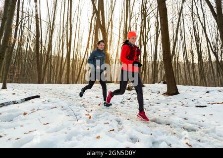 Man and Woman Running Together Through Woods in Winter. Smiling Couple Running Along Trail Through Forest on Cold Winter Day. Stock Photo