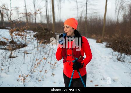 Female Runner Drinking Water from Soft Flask in Woods on Winter Day. Woman Wearing Hydration Vest Taking Break From Trail Running in Woods and Sipping Stock Photo