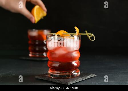 Woman squeezing fresh orange in glass with tasty Negroni cocktail on dark background Stock Photo