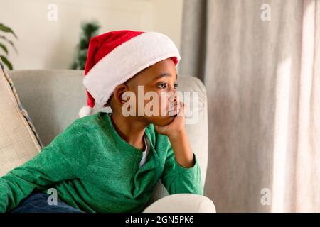 Thoughtful african american boy wearing santa hat and sitting in armchair at christmas time Stock Photo