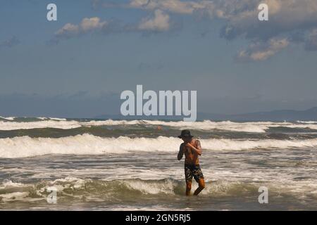 Tanned and tattooed young man standing on the shore of the rough sea with big waves and the Tuscan coastline in the background, Livorno prov., Tuscany Stock Photo