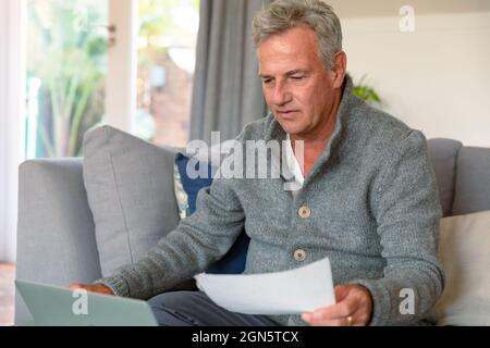 Focused caucasian senior man sitting on sofa, doing paperwork, using laptop Stock Photo