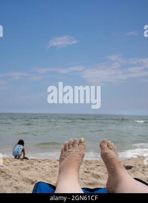 barefeet on a beach chair facing the ocean sea water shore line with daughter playing in the sand Stock Photo