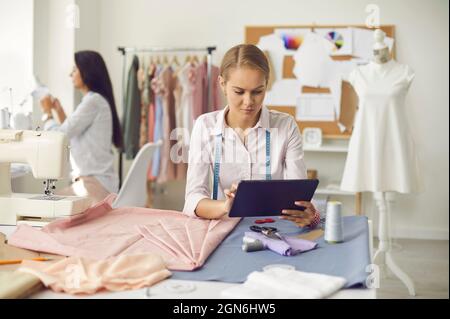 Female dressmaker sitting at table in her sewing atelier and using modern tablet Stock Photo