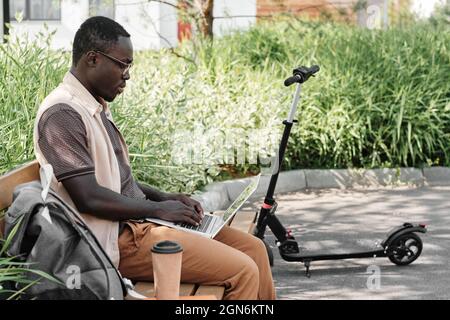 Horizontal medium side view shot of young African American man wearing casual outfit sitting on bench outdoors texting something on his laptop Stock Photo