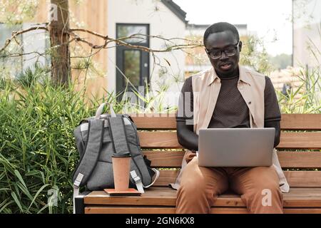 Horizontal medium portrait of handsome young African American man wearing casual outfit sitting on bench outdoors doing some freelance work using his laptop Stock Photo