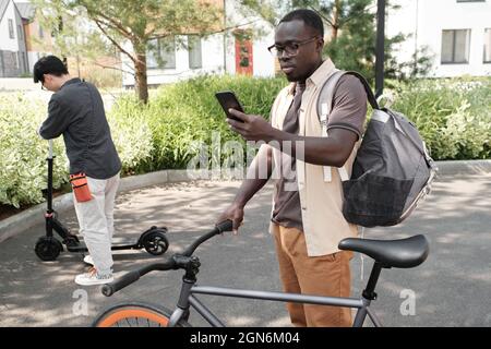 Horizontal shot of young African American man standing outdoors checking something on his smartphone while parking his bicycle Stock Photo