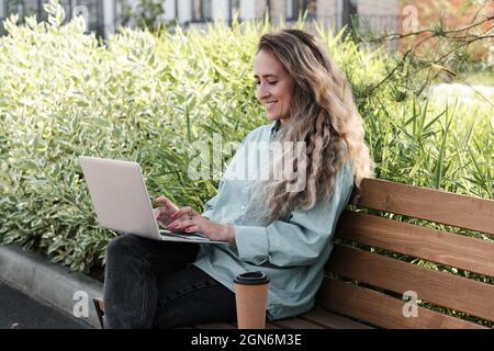 Horizontal medium portrait of young adult woman with long curly hair sitting on bench in city park working remotely on laptop Stock Photo