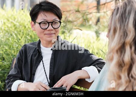 Portrait of young Asian woman with short hair wearing eyeglasses spending time outdoors with her friend drinking coffee and chatting Stock Photo