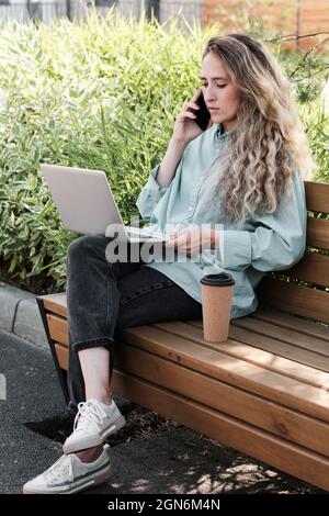 Vertical full portrait shot of stylish young woman with long curly hair sitting on bench in city park working remotely on laptop Stock Photo