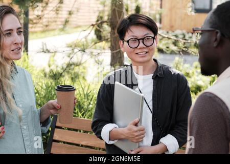 Horizontal medium shot of three young multi-ethnic colleagues standing together outdoors on summer day talking about something during coffee break Stock Photo