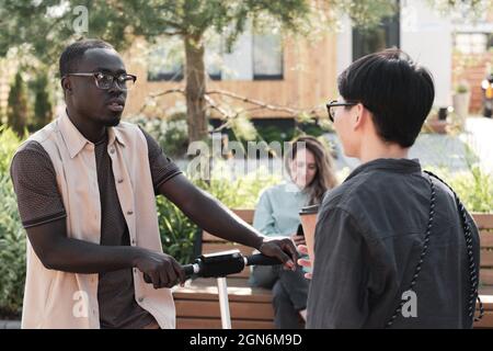 Horizontal medium portrait of young African American man and Asian woman running into each other outdoors standing in park and having small conversation Stock Photo