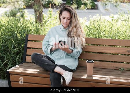 Horizontal medium long portrait of attractive Caucasian woman with long hair wearing casual outfit sitting on wooden bench outdoors using smartphone Stock Photo