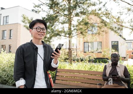 Horizontal medium shot of young Asian woman with short hair wearing eyeglasses spending time outdoors walking and recording voice message using smartphone Stock Photo
