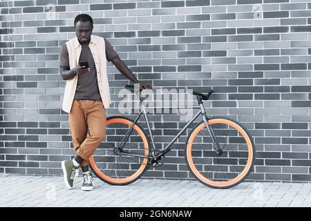 Horizontal long shot of young Black man wearing casual clothes standing with bicycle against gray brick building watching something on smartphone, copy space Stock Photo