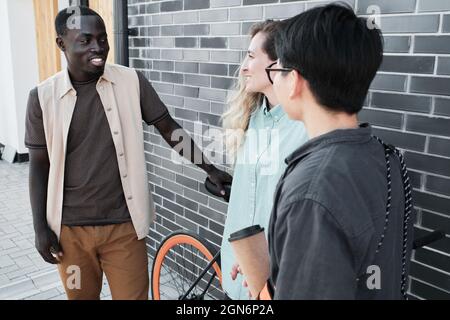 Portrait of young African American man and his female colleagues wearing casual clothes standing together outdoors chatting about various things during coffee break time Stock Photo