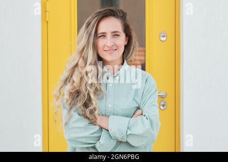 Horizontal waist up portrait shot of attractive young adult Caucasian woman with long wavy hair wearing pale green shirt standing with arms crossed against yellow door, looking at camera Stock Photo