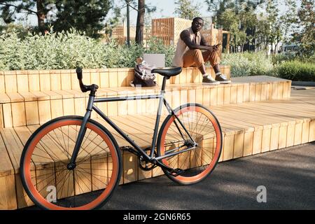 Horizontal selective focus shot of young African American man sitting on bench in modern city park looking at his bicycle parked nearby Stock Photo