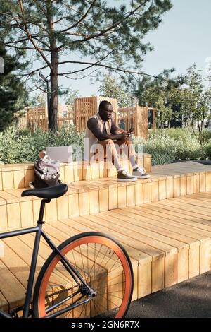 Vertical long shot of young Black man wearing casual outfit sitting on bench in urban park on sunny summer day watching something on smartphone Stock Photo