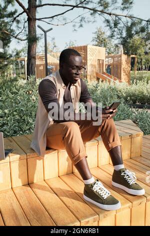 Vertical full shot portrait of handsome young Black man wearing casual outfit sitting in urban park watching something in Internet on his smartphone Stock Photo