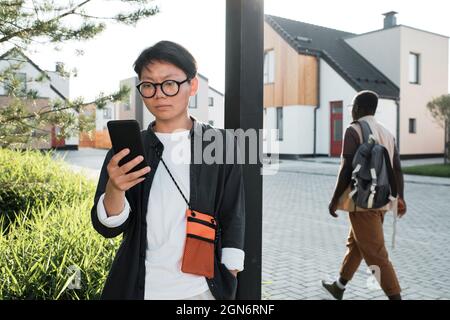 Horizontal medium portrait of modern young Asian woman wearing casual clothes and eyeglasses standing outdoors checking data on her smartphone Stock Photo