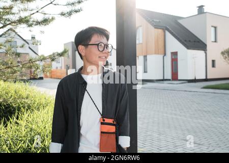 Horizontal medium portrait shot of young Asian woman wearing stylish casual outfit with eyeglasses standing outdoors looking away smiling, copy space Stock Photo