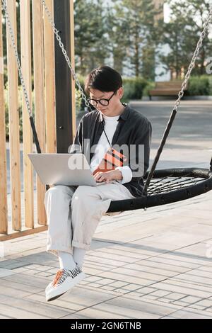 Vertical shot of stylish young Asian woman wearing casual outfit spending time outdoors sitting on swing in urban park doing remote work on laptop Stock Photo